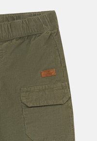Pantalon en velours côtelé vert avec une taille élastique et une poche latérale, comportant une petite étiquette de marque en cuir marron sur le tissu.