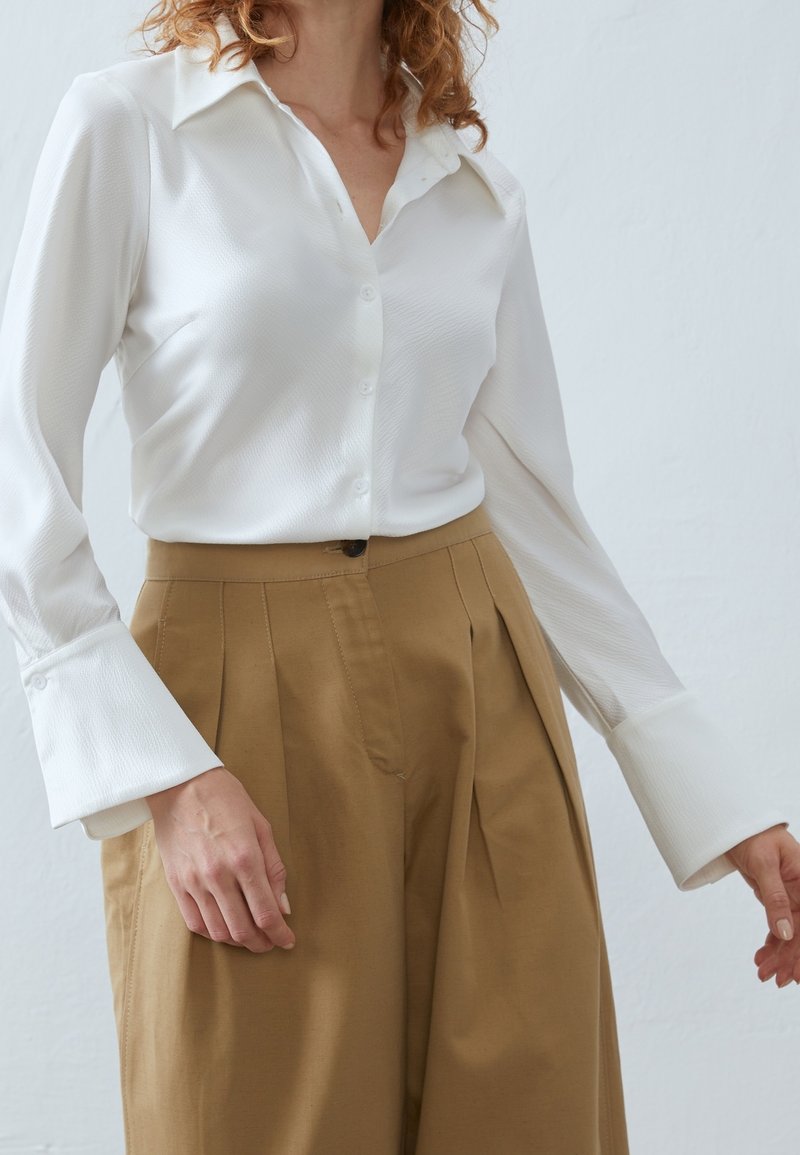 Femme portant une chemise blanche à boutons avec de larges poignets et un pantalon marron ample taille haute sur un fond uni.