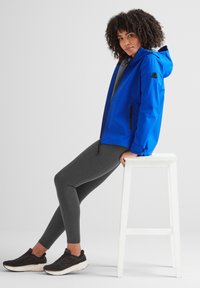 Woman with curly hair wearing a blue hooded jacket, gray leggings, and black sneakers, sitting on a white wooden stool.