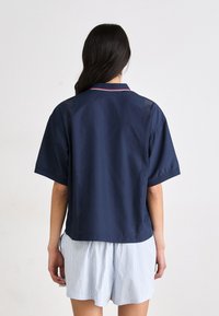 Camicia a maniche corte blu navy con colletto, dotata di pannelli ventilati sulla schiena e accenti rossi a contrasto. Abbinata a pantaloni corti a righe.