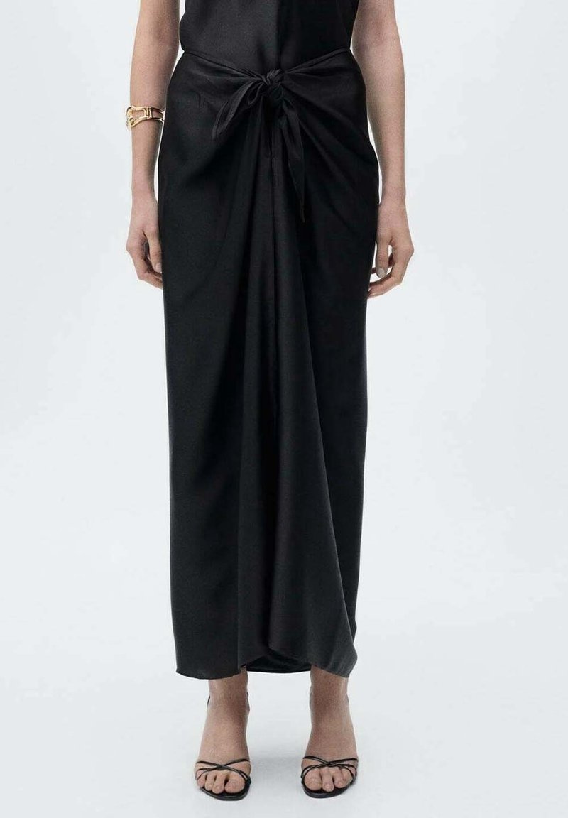 Femme portant une longue robe noire nouée à la taille, avec des sandales noires à brides et un bracelet en or au poignet gauche.
