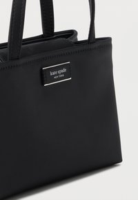 Μαύρη τσάντα Kate Spade New York με δύο λαβές και ορθογώνια ετικέτα με το λογότυπο μπροστά, πάνω σε μονόχρωμο φόντο.