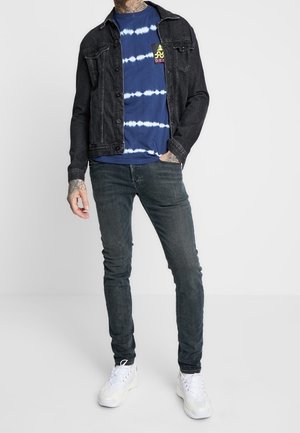 Černá džínová bunda přes námořnicky modré tričko s batikovaným vzorem a bílými motivy. Úzké tmavé džíny a bílé tenisky dokončují outfit.