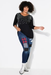 Haut noir avec des détails en étoile argentée, décolleté en V et manches trois-quarts. Jeans bleus déchirés avec des pièces en patchwork de couleurs et textures variées.
