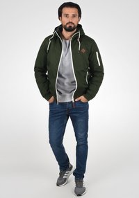 Zöld nylon dzseki kapucnival, fehér cipzárral és zsebekkel. Szürke pulóver van alatta, kék farmer és szürke sportcipő fehér csíkokkal.