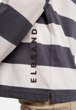 Schwarz-weiß gestreiftes, horizontal verlaufendes Hemd mit der vertikal nahe am Saum gedruckten Aufschrift „ELBSAND“, getragen über beigen, gerippten Hosen.