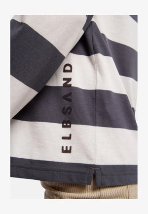 Schwarz-weiß gestreiftes, horizontal verlaufendes Hemd mit der vertikal nahe am Saum gedruckten Aufschrift „ELBSAND“, getragen über beigen, gerippten Hosen.