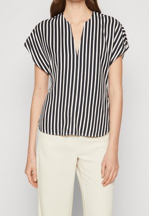 Femme portant un chemisier rayé vertical noir et blanc à manches courtes et un pantalon taille haute beige clair sur un fond uni.