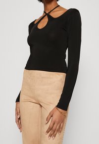 Maglietta nera a maniche lunghe con scollatura a oblò e una vestibilità aderente. Il materiale sembra liscio con una leggera lucentezza, in contrasto con pantaloni beige.
