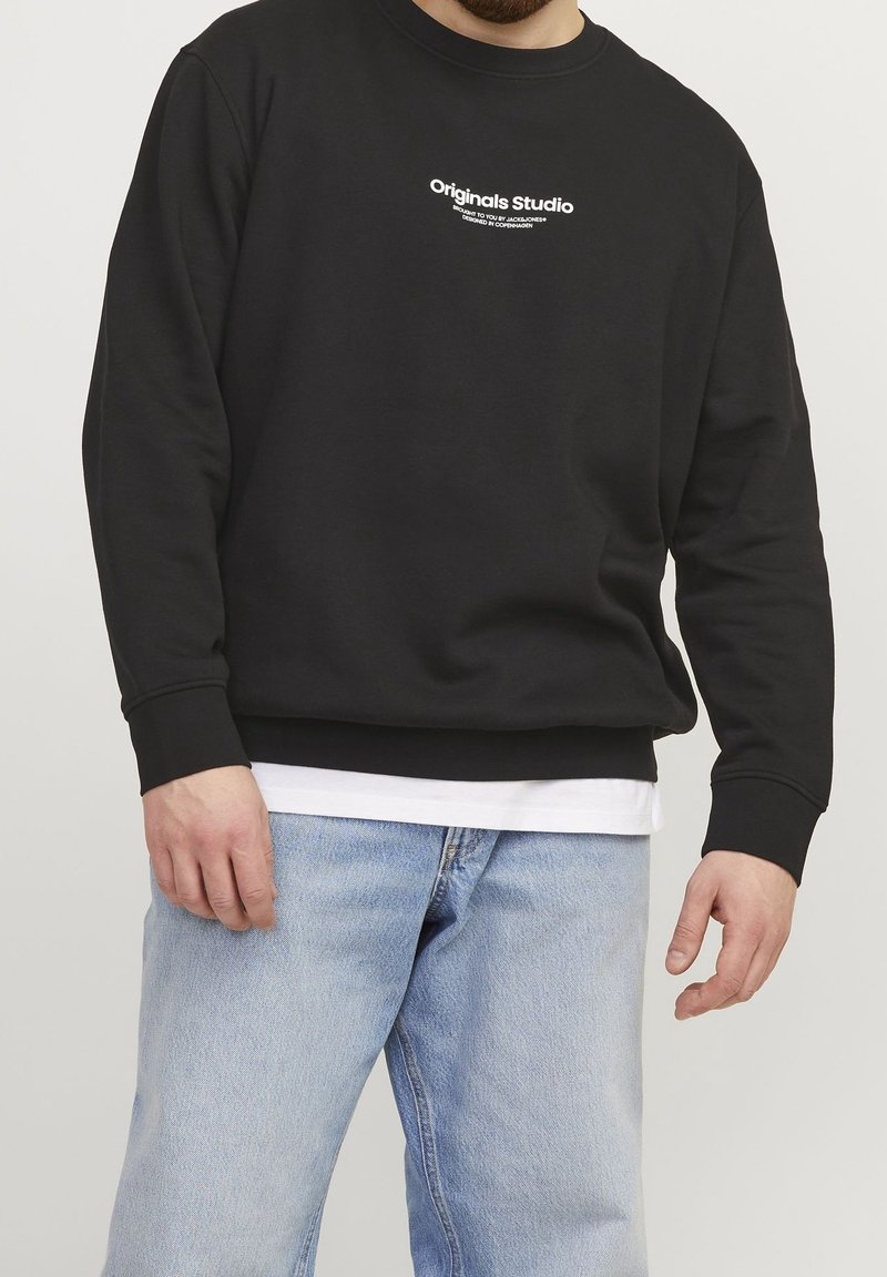 Felpa nera con collo tondo, maniche lunghe e polsini a coste. Presenta la scritta bianca "Originals Studio" sul petto; indossata con jeans azzurri chiari.