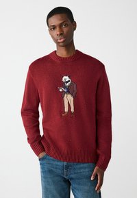 Czerwony sweter dzianinowy z prążkowanym dekoltem i mankietami, z haftowaną postacią borsuka w marynarce i beżowych spodniach trzymającego notes.