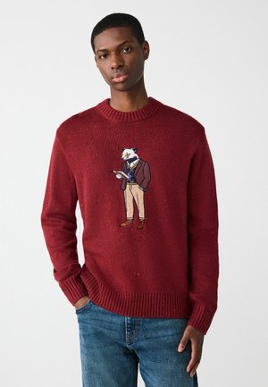 Raudona megzta striukė su raštuotu apykakle ir rankovėmis, su siuvinėtu šernu, vilkinčiu švarką ir smėlio spalvos kelnes, laikantį užrašų knygelę.