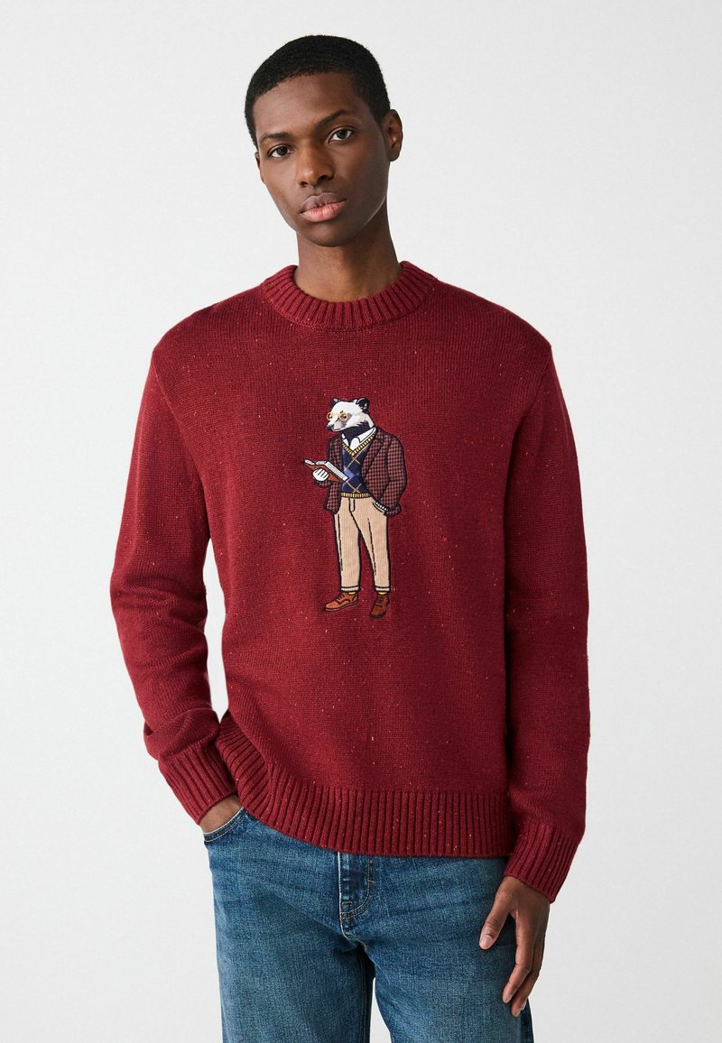 Czerwony sweter dzianinowy z prążkowanym dekoltem i mankietami, z haftowaną postacią borsuka w marynarce i beżowych spodniach trzymającego notes.