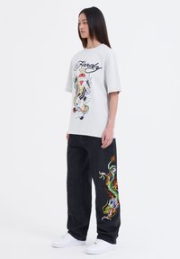 Grijze oversized t-shirt met een kleurrijke drakenafbeelding, gecombineerd met zwarte jeans met een opvallende drakenborduring aan de zijkant. Witte sneakers.