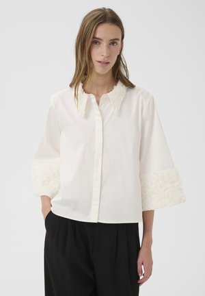 Femme portant une blouse blanche avec des poignets et un col texturés, associée à un pantalon noir ample, se tenant devant un fond uni.
