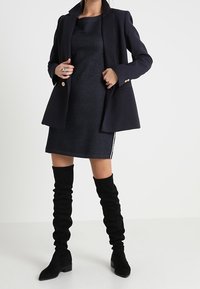 Blazer en velours côtelé marine sur une robe sombre, accompagné de bottes en tissu noires montantes. Le blazer possède deux boutons et des poches.
