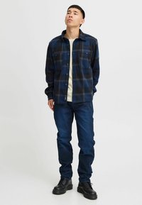 Flanellskjorte i marineblå og svart rutete, knapper foran, med to brystlommer. Fitted blå jeans og svarte snørebåndsstøvler. Nøytral bakgrunn.