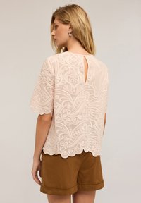 Donna che indossa una blusa di pizzo rosa chiaro con motivi intricati e bordi scallop, abbinata a pantaloni corti marroni con risvolto, di spalle.