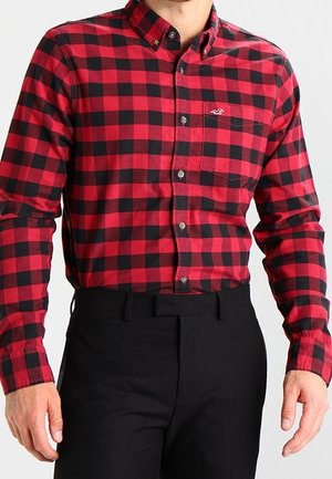 Man die een rood-zwart geruit overhemd met lange mouwen draagt, ingestopt in een zwarte broek, tegen een effen achtergrond.