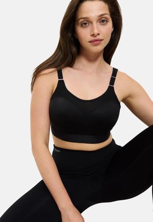 Joven mujer con cabello largo y castaño, vestida con un sujetador deportivo negro y leggings negros, sentada con una rodilla levantada sobre un fondo blanco.