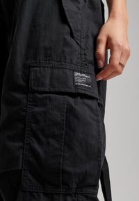 Czarne spodnie cargo z boczną kieszenią i etykietą pielęgnacyjną wszytą na udzie. Tkanina ma gładką, matową teksturę.