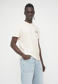 Ανοιχτό μπεζ βαμβακερό μπλουζάκι με κοντά μανίκια και στρογγυλή λαιμόκοψη, με ένα μικρό πράσινο και λευκό λογότυπο στο στήθος. Φορεμένο με ανοιχτό μπλε τζιν.