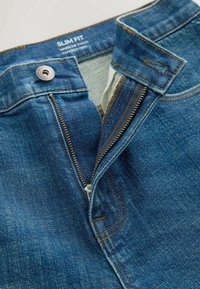 Jean slim bleu avec fermeture éclair métallique partiellement dézippée et bouton, montrant une cuisse fine et un ourlet effilé, étiquette à l'intérieur de la ceinture.