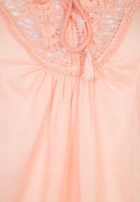 Peach-colored garment with a crocheted lace panel at the neck, featuring a gathered texture and drawstring with tassels.