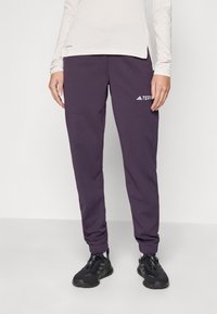 Lila mjukisbyxor med elastiska muddar, slät textur, två sidofickor och en vit logotyp på låret. Svarta skor med snörning.