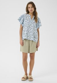 Modrá blúzka s kvetinovým vzorom, krátkymi volánovými rukávmi a výstrihom do V, kombinovaná s svetlo béžovými šortkami a béžovými šľapkami.