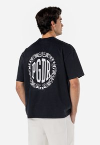 Must t-särk lühikeste varrukatega, valmistatud puuvillast, millel on valge ornate ringikujuline disain ja tagaküljel tekst "PGDR". Lihtne, mugav lõige.