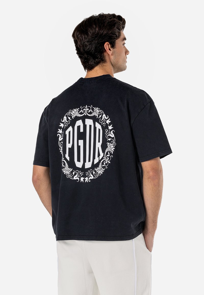 Must t-särk lühikeste varrukatega, valmistatud puuvillast, millel on valge ornate ringikujuline disain ja tagaküljel tekst "PGDR". Lihtne, mugav lõige.