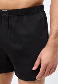 Gros plan sur une personne portant un short noir à taille élastique avec une petite étiquette rectangulaire sur la ceinture, la main reposant sur le côté.