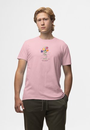Jonge man met lichtbruin haar die een roze T-shirt draagt met een kleurrijk bloemmotief en de tekst "Picasso", en donkergroene broek.
