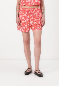 Røde blomstertrykte shorts med elastisk midje og snor. Brukt med svarte spisse flats. En enkel hvit bakgrunn.