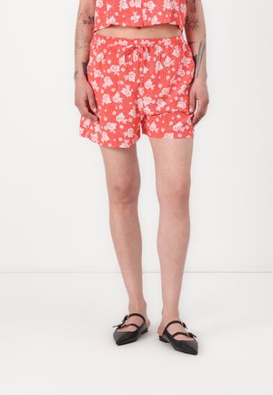 Rode bloemenprint shorts met een elastische tailleband en koord. G gedragen met zwarte puntige ballerina's. Effen witte achtergrond.