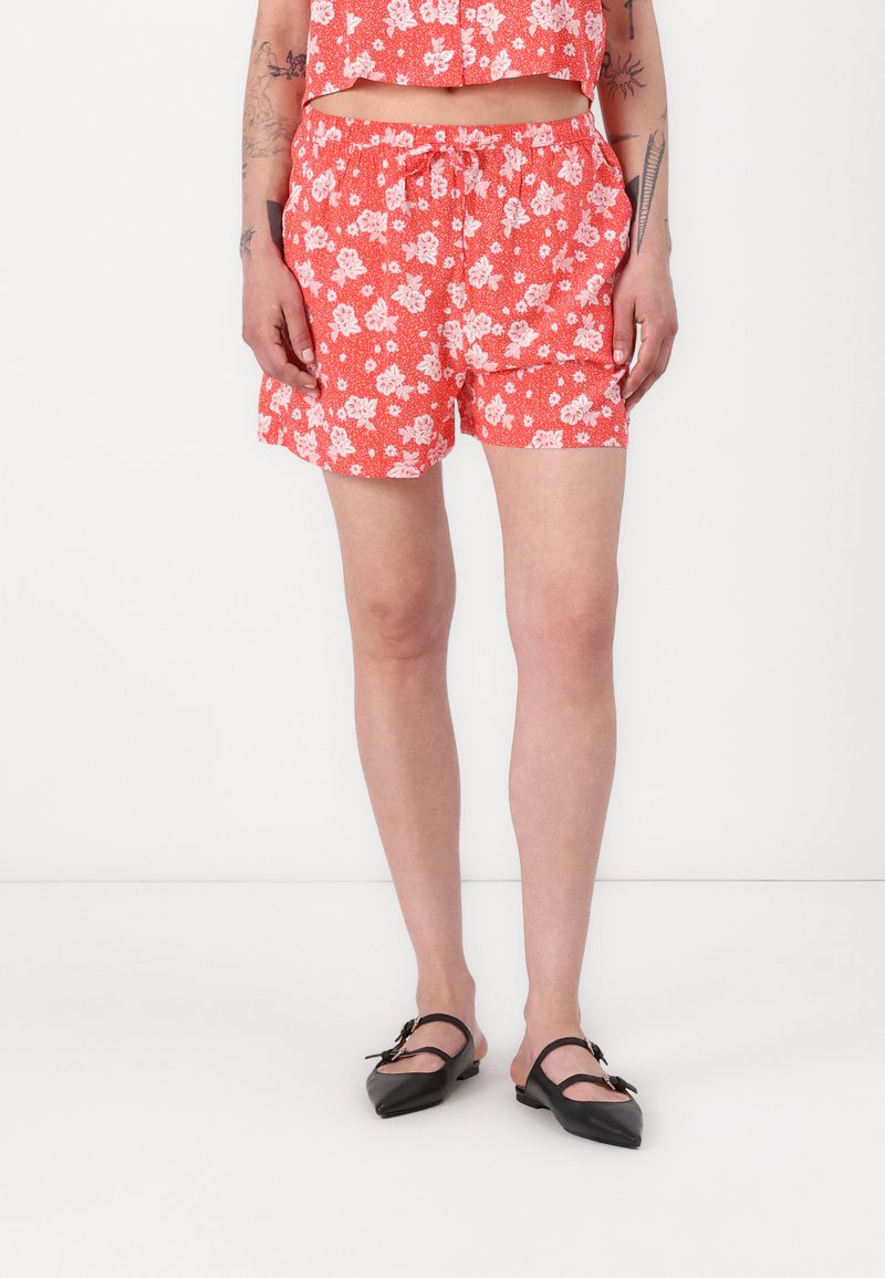 Røde blomstertrykte shorts med elastisk midje og snor. Brukt med svarte spisse flats. En enkel hvit bakgrunn.
