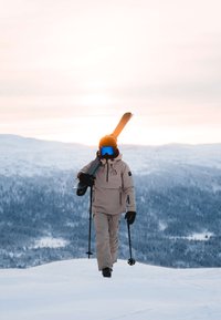 Snowboardåkare klädd i en ljusbeige jacka och byxor, bär på skidor, med orange hjälm och blå goggles, på ett snötäckt bergslandskap.