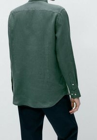 Camicia verde a maniche lunghe in un tessuto morbido con taglio dritto. Presenta un colletto, polsini con bottoni e un orlo basso con bordi arrotondati.