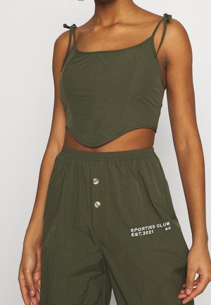 Kobieta nosząca oliwkowozielony krótki top na ramiączkach wiązanych na kokardkę oraz pasujące wysokie spodnie z napisem "Sporting Club Est. 2021".