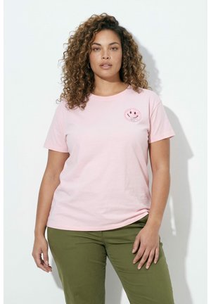 Vrouw met krullend haar draagt een lichtroze t-shirt met een kleine patch van een glimlachend gezicht en olijfgroene broek, staand tegen een witte achtergrond.