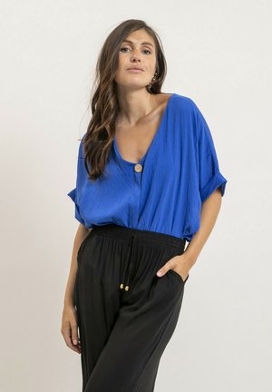 Femme aux longs cheveux bruns, vêtue d'un haut bleu ample boutonné et d'un pantalon noir à cordon, posant avec une main dans la poche sur un fond uni.