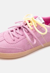 Ροζ σουέντ αθλητικό παπούτσι με μπεζ σόλα, διαθέτει δύο δεμένα κορδόνια—ένα ροζ και ένα κίτρινο—διακοσμητικές ραφές και εμφανές ετικέτα στη γλώσσα του παπουτσιού.