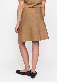 Brun A-linje kjol i texturerad tyg, som faller ovanför knät, i kombination med svarta ballerinaskor med rosettdekoration. Sidogångdragkedja synlig.