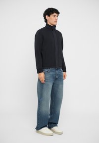 Veste zippée noire avec un col montant, en matériau doux, et deux poches avant, associée à un jean bleu ample et des baskets blanches.