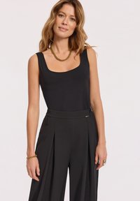 Kobieta z falującymi włosami, ubrana w bezrękawnik czarny top, czarne spodnie z wysokim stanem, złoty naszyjnik łańcuszkowy, bransoletkę i pierścionek, stoi na tle białym.