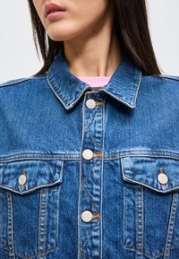 Gros plan d'une personne portant une veste en jean bleue avec des boutons métalliques et deux poches poitrine, montrant une partie de son visage et de son cou.