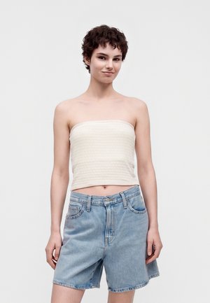 Jeune personne aux cheveux courts et bouclés portant un haut blanc sans bretelles et un short en jean bleu clair ample, debout devant un fond uni.