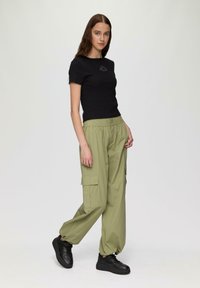 Zwarte top met korte mouwen en borduursel aan de voorkant, gecombineerd met loszittende olijfgroene cargobroek met zijzakken en een elastische tailleband. Zwarte sneakers.