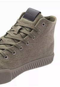 Zapatilla alta de lona verde con suela de goma, que presenta agujetas a juego, dos ojales de metal y una etiqueta negra con logo en el tobillo.